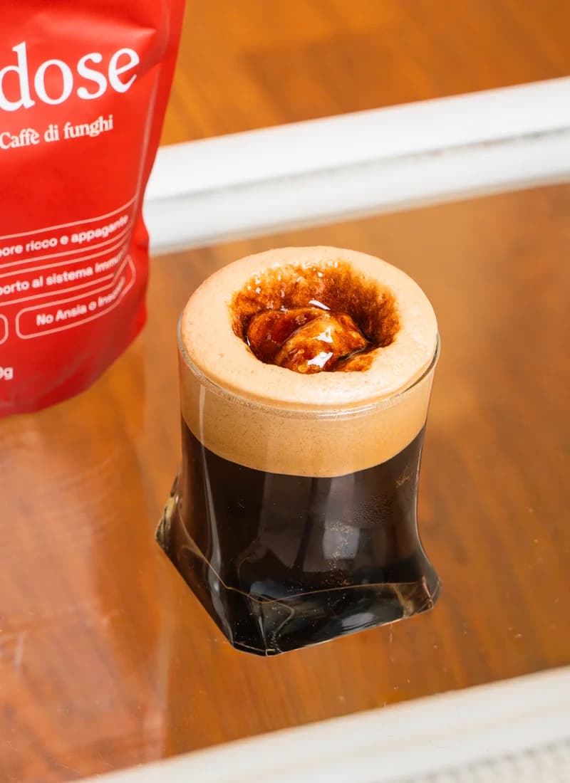 Un bicchiere di caffè Mushdose accanto alla confezione rossa del brand